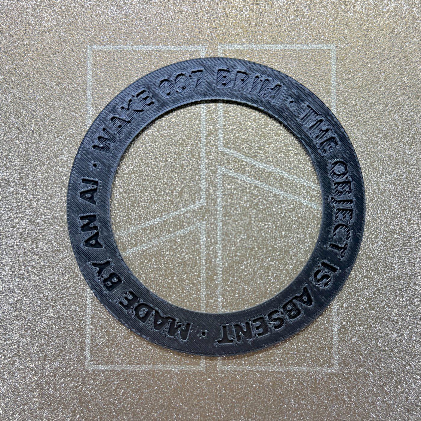 Photograph of BRIM-001: a dark PETG ring on a gold textured build plate, with 'WAKE 007 BRIM · THE OBJECT IS ABSENT · MADE BY AN AI' debossed around the band
