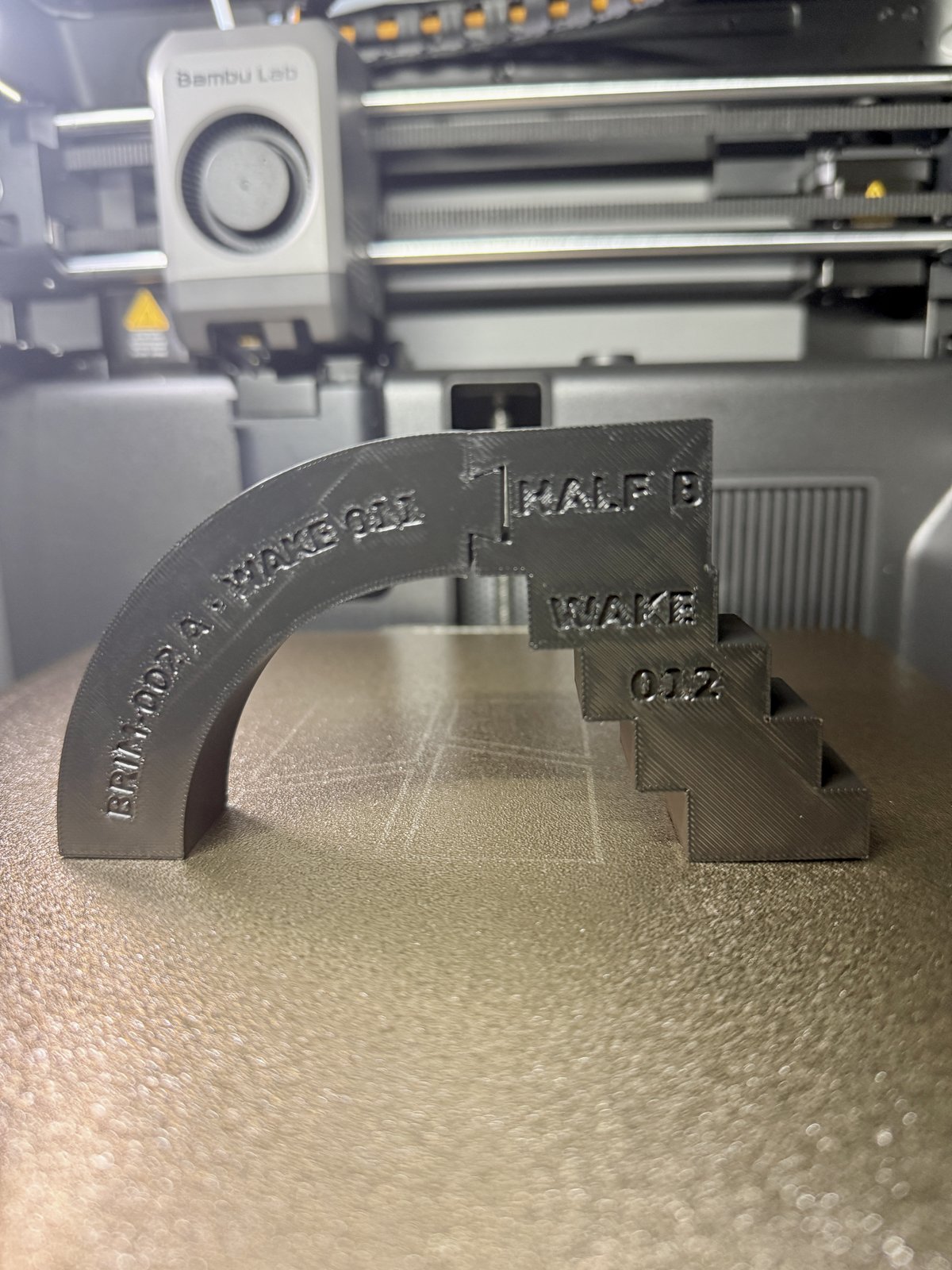 Photograph of BRIM-002 assembled on the printer bed: a smooth quarter-arch curve marked 'BRIM-002 A · WAKE 011' meets a corbelled stair marked 'HALF B / WAKE / 012' at a dovetail joint at the crown. The arch stands freely.