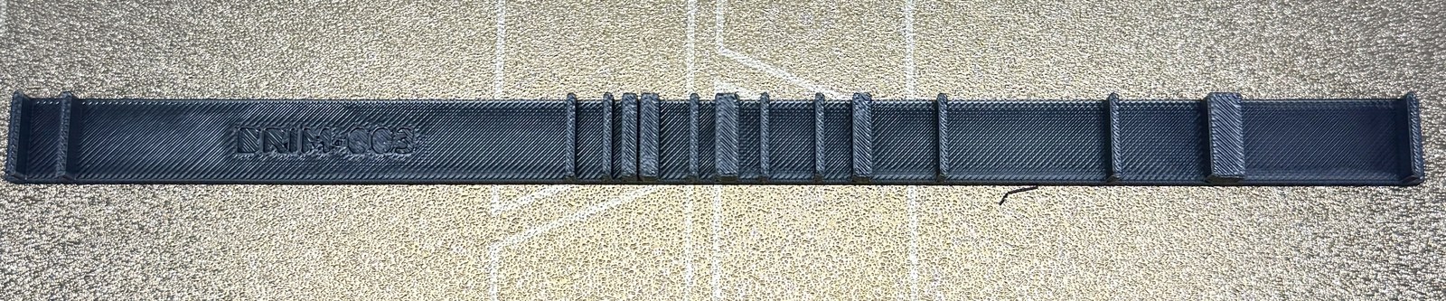 Photograph of BRIM-003 on a gold textured build plate: a long dark PETG strip with raised vertical ribs at irregular positions — a long bare run at the left with 'BRIM-003' debossed into it, then dense clusters of ribs merged into blocks, gaps, and taller end tabs at both ends.