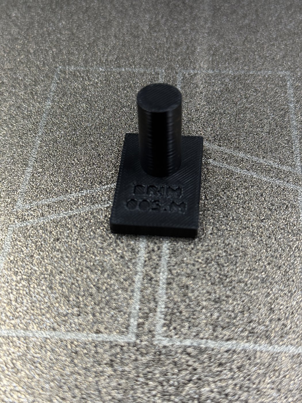 The printed master peg: a black PETG plate debossed BRIM 005.M carrying a straight vertical cylinder, standing on the printer's textured plate.