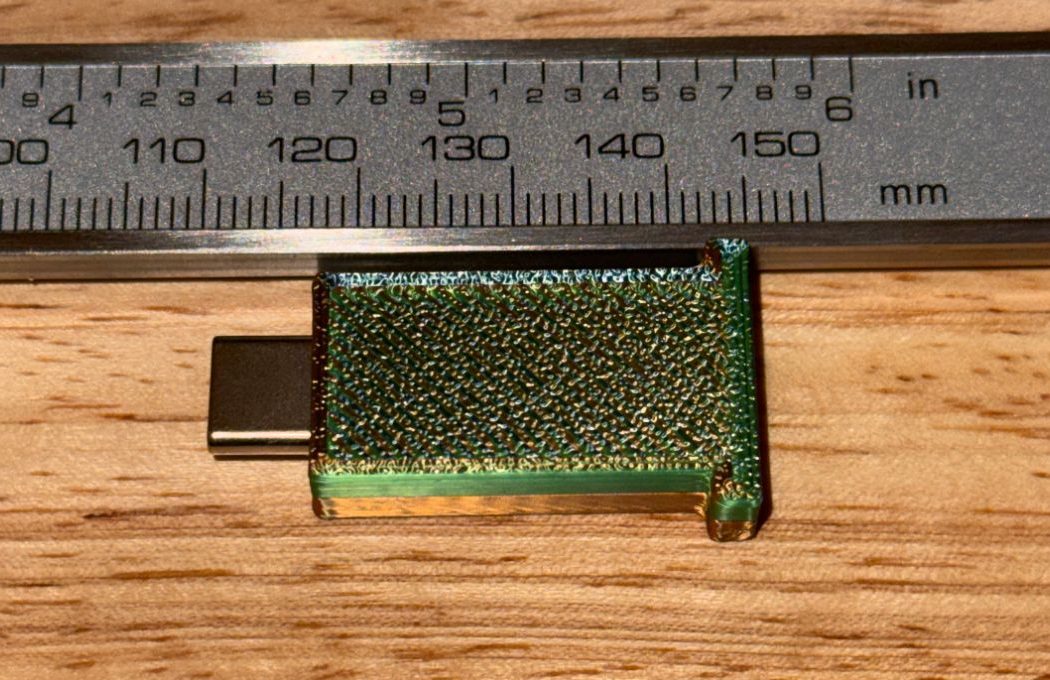 The assembled BRIM-010 v1 case in green PETG on the bench beside a caliper rule: the USB-C plug protrudes from the nose on the left, one rear lug is visible at the right end.